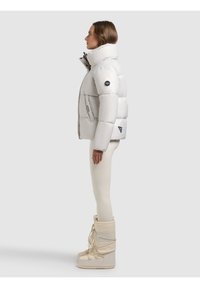 Donna con giacca imbottita bianca, leggings color crema e stivali invernali beige, in piedi di profilo su uno sfondo grigio chiaro.