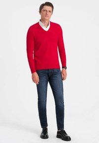 Czerwony sweter z dzianiny z dekoltem w serek, noszony na białej koszuli, parowany z ciemnoniebieskimi dżinsami i czarnymi wiązanymi butami.
