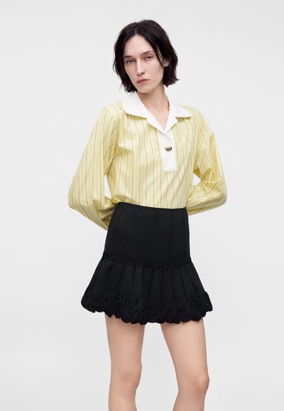 Femme aux cheveux courts et foncés portant un chemisier jaune à rayures avec un col blanc et une mini-jupe noire à volants, debout, les mains derrière le dos.