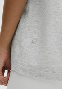 T-shirt en coton gris clair avec un ourlet arrondi, présentant un petit logo brodé en bas à gauche. Texture douce et design décontracté.