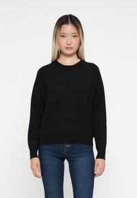 Fekete kötött pulóver kerek nyakkal, ejtett vállakkal és hosszú ujjal. Simább tapintás, laza fazon, kék farmernadrággal párosítva.