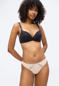 Γυναίκα με σγουρά μαλλιά που φοράει μαύρο σουτιέν με λουράκια λογότυπου και μπεζ εσώρουχο στυλ μπικίνι με δαντέλα, στέκεται μπροστά σε ένα απλό φόντο.