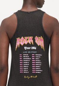Juodas marškinėlis be rankovių su užrašu "ROCK ON" rožinės ir geltonos spalvos. Dizainas apima turo datas rožine ir balta spalvomis. Tekstūra su apipjaustytu kraštu.