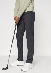 Tumši pelēki golfa bikses, kas izgatavotas no viegla auduma, ar tievu piegriezumu. Persona tur golfa nūju, valkājot baltus sporta apavus.