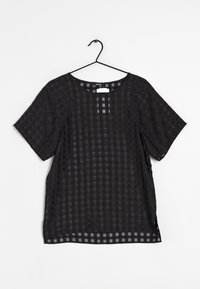 Blouse noire à manches courtes avec motif à carreaux transparent, suspendue sur un cintre noir contre un fond blanc.