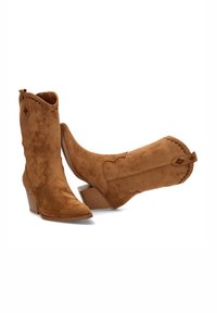 Botas vaqueras de ante en color tostado con tacón apilado, punta afilada, costuras decorativas y lengüetas en la parte superior para facilitar su uso.