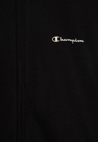 Čierna mikina na zips vyrobená z kombinácie bavlny. Má malé biele logo "Champion" na ľavej strane hrude a rebrovanú textúru.