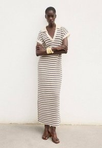 Striped, long knit dress in cream and navy, featuring a V-neck, short sleeves, and side slits. Worn with brown heeled sandals and yellow bracelets.
