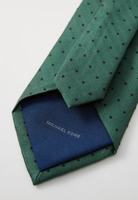 Cravate en chevron vert avec des pois noirs, pliée, révélant une doublure bleue marine étiquetée "MICHAEL KORS." Texture lisse et design moderne.