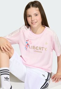 Jong meisje zittend, draagt een roze Adidas Liberty London-shirt, witte Adidas short en witte sokken met zwarte strepen.