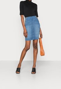Falda lápiz de mezclilla con cintura alta, lavado claro, con botones laterales. Combinada con una blusa negra y sandalias negras de punta abierta.