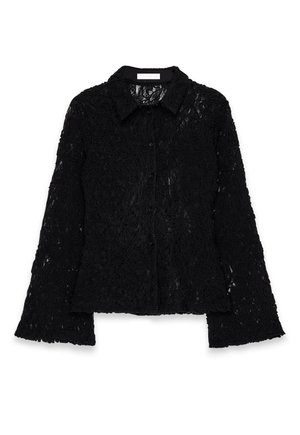 Mujer vestida con una blusa negra de encaje con botones, pantalones negros holgados, zapatos color burdeos, sosteniendo un bolso negro, de pie junto a unos escalones de piedra blanca y una columna.