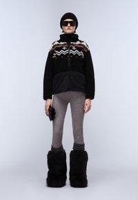 Persona che indossa un berretto nero, grandi occhiali da sole, una giacca in pile nera a fantasia, leggings grigi e stivali alti neri di pelliccia, mentre tiene dei guanti neri.