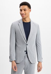 Grau-weiß gestreifter Blazer mit zwei Fronttaschen, einem einzigen Knopfverschluss und Rückenschößchen. Getragen über einem marineblauen T-Shirt.