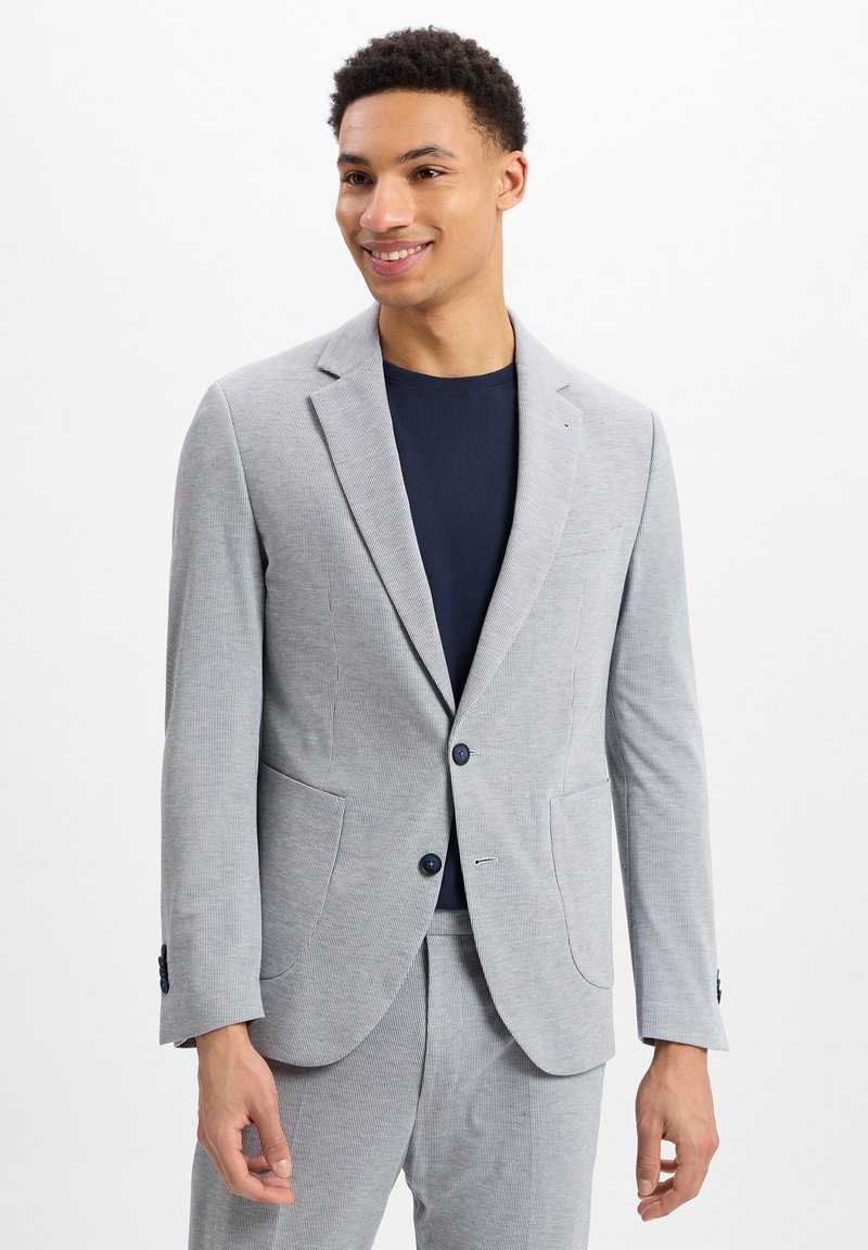 Grau-weiß gestreifter Blazer mit zwei Fronttaschen, einem einzigen Knopfverschluss und Rückenschößchen. Getragen über einem marineblauen T-Shirt.