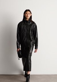 Crna kožna jakna s visokim ovratnikom i kopčanjem na snap, uparena s odgovarajućom suknjom. Glatka tekstura, prilagođen dizajn i elegantna silueta.