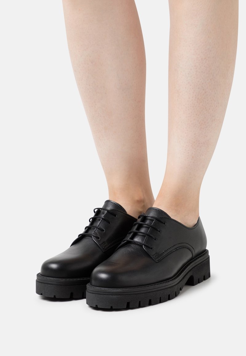 Chaussures derby en cuir noir à lacets avec semelles épaisses et robustes portées sur des jambes nues sur un fond clair uni.