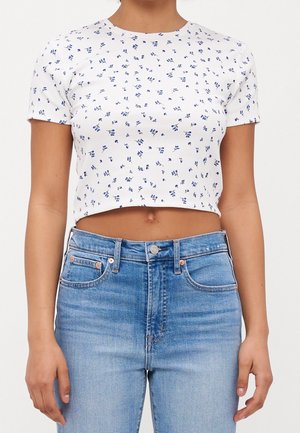 Kobieta ubrana w biały krótki t-shirt z małymi niebieskimi kwiatowymi wzorami oraz jasnoniebieskie jeansy z wysokim stanem na jednolitym tle.