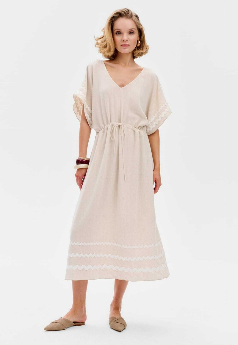 Beige linnen jurk met een v-hals, een trekkoord in de taille en brede mouwen. Heeft witte zigzag borduursels aan de onderkant. Draag het met beige instapschoenen.