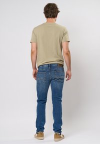 Camiseta de cuello redondo beige, jeans azul slim-fit y zapatillas color marrón claro. El denim tiene bolsillos traseros con detalles de costura. Textura suave.