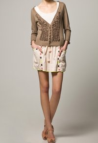 Brązowy kardigan z falbankami, zestawiony z jasną spódnicą z dekoracyjnymi kieszeniami i neonowymi lamówkami. Dopełnieniem stroju są płaskie sandały.