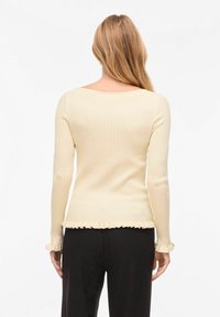 Femme aux longs cheveux blonds portant un haut crème à manches longues côtelé avec ourlet et poignets à volants, associée à un pantalon noir, vue de dos.