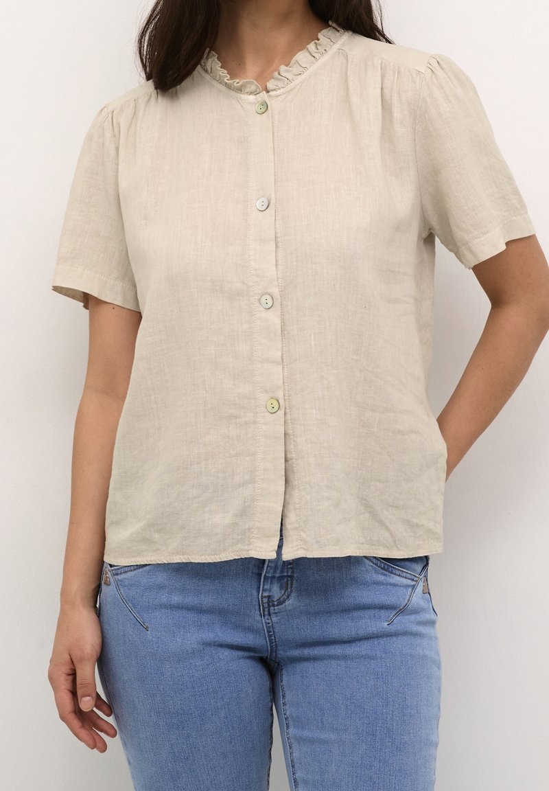 Camicia beige in lino con maniche corte, collo arricciato e tre bottoni chiari sul davanti, abbinata a jeans blu. Design semplice e con texture.