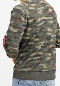 Sudadera de camuflaje en verde, marrón y negro con puños y dobladillo acanalados. Presenta un bolsillo lateral con una etiqueta de tela roja adjunta.