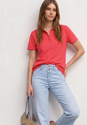 Jeune femme en polo corail à manches courtes et jean bleu clair, debout, tenant un sac cabas tissé avec des anses bleues devant un mur blanc.