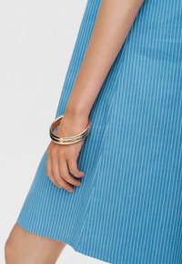 Modra črtasta obleka s razširjenim robom. Roka drži tri zlate zapestnice. Tkanina se zdi gladka z navpičnimi belimi črtami.