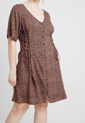 Woman wearing a knee-length, short-sleeve, V-neck dress with a small orange, blue, and black floral pattern and a side drawstring.