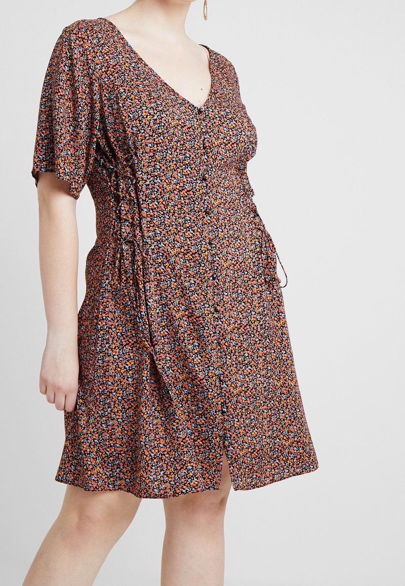 Femme portant une robe mi-longue à manches courtes et col en V, avec un petit motif floral orange, bleu et noir, et un cordon de serrage sur le côté.