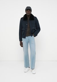 Marineblauwe jas met zwarte bontkraag, bruine trui, lichtblauwe wijde spijkerbroek en witte sneakers, gedragen met een grijze pet.