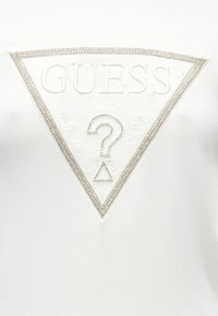 Λευκό φούτερ με υφασμένο τριγωνικό λογότυπο, όπου η λέξη «GUESS» εμφανίζεται σε ανάγλυφα γράμματα με ασημένιες διακοσμητικές πέτρες.