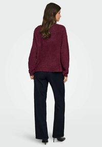 Burgunderfarbener strukturierter Pullover mit langen Ärmeln, lässiger Passform und Rundhalsausschnitt, kombiniert mit dunkelblauen weit geschnittenen Hosen und schwarzen High Heels.