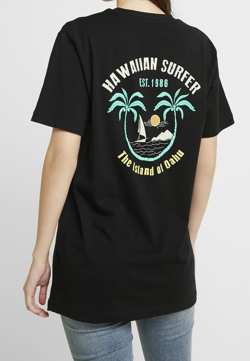 Černé bavlněné tričko s potiskem na zádech, který zobrazuje palmy, plachetnici, slunce a text "HAWAIIAN SURFER EST. 1986" a "Ostrov Oahu."