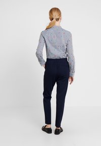 Kvinna med blont hästsvans iförd en randig skjorta med färgglada mönster, marinblå byxor med smal passform och svarta loafers som står mot en vit bakgrund.