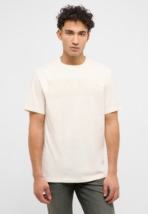 Junger Mann mit lockigem Haar, der ein einfaches hellbeiges T-Shirt mit kurzen Ärmeln und dunkle Hosen trägt, vor einem weißen Hintergrund stehend.