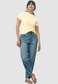 Gelbes Baumwoll-T-Shirt mit floraler Stickerei auf der Brust, kombiniert mit hochgeschnittenen blauen Jeans. Das Model trägt hellbraune Ballerinas.