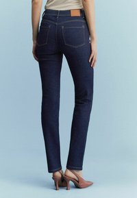 Žena v tmavě modrých slim-fit džínách a světle růžových lodičkách na podpatku, stojící na světle modrém pozadí.