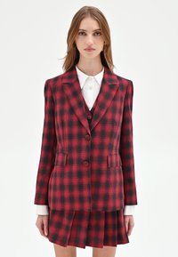 Blazer a quadri rossi e neri con bavero a lancia, chiusura con doppio bottone e tasche frontali, abbinato a una gonna plissettata coordinata.