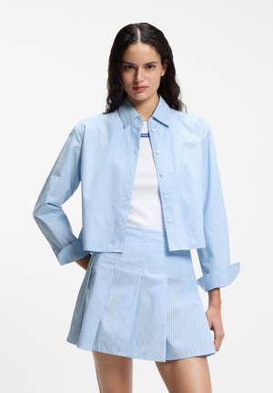 Kobieta ubrana w jasnoniebieską, prążkowaną, krótko skrojoną koszulę z guzikami oraz dopasowaną plisowaną spódnicę, na białej bluzce, stojąca na jednolitym tle.