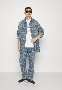 Traper jakna i hlače s grafitnim uzorkom u plavoj boji, u kombinaciji s bijelom majicom i crnim sportskim cipelama. Model nosi sunčane naočale.