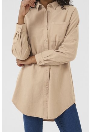 Personne portant une chemise beige à manches longues boutonnée avec une poche poitrine et un jean bleu, posant avec une main près du visage.