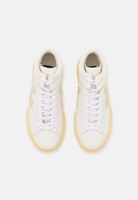 White high-top sneakers made of smooth leather, featuring a textured cream midsole, padded collar, white laces, and yellow rubber outsole.
