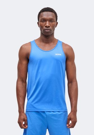 Hombre atlético que lleva una camiseta sin mangas azul de Borg y pantalones cortos a juego, de pie y mirando hacia adelante sobre un fondo liso y claro.