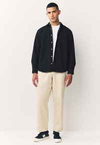 Schwarzes Hemd zum Knöpfen über einem weißen T-Shirt, kombiniert mit cremefarbenen Hosen und dunklen Turnschuhen. Das Hemd hat eine Tasche und lange Ärmel.