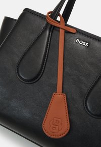 Svart läderhandväska med subtil matt textur, framträdande sömmar, en beige läderdetalj och silverfärgad "BOSS"-logotyp.