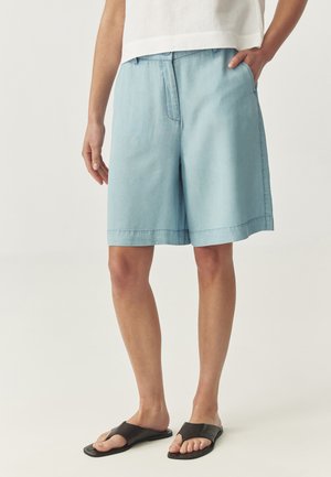 Short bleu clair arrivant au genou sur une personne portant un haut blanc et des sandales noires à bout ouvert, debout avec une main dans la poche.