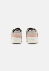 Les baskets présentent un mélange de suède rose pâle et de cuir beige lisse, avec une semelle blanche et verte. Le patch texturé au talon affiche le logo "LACOSTE".